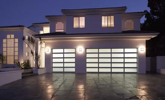 Aluminum garage doors on a large two-story home at dusk, with lights on inside, visible through garage door blurred windows.